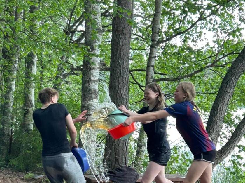 Kolme nuorta rippileirillä leikkimässä vesisotaa lammen rannalla.