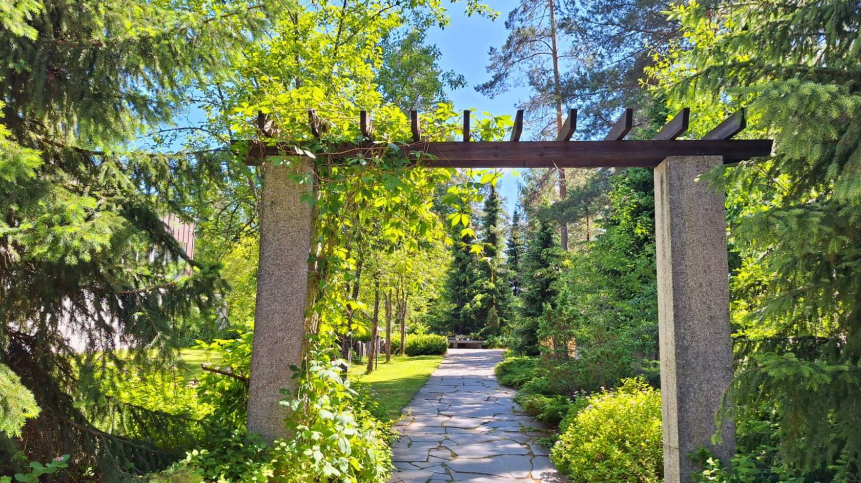 Luonnonkivillä päällystetty, puiden ja pensaiden reunustama polku johtaa kivestä ja puusta tehdyn portin läpi.