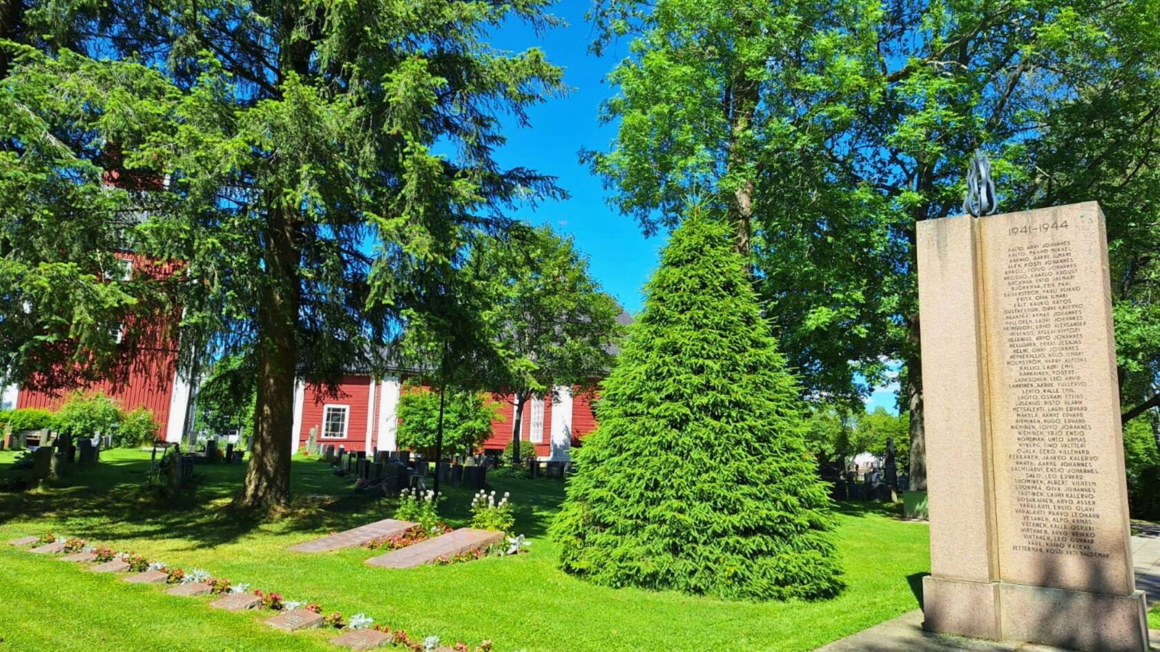 Aurinkoinen kesäpäivä, etualalla sankarihautoja ja muistomerkki, kauempana hautausmaan puita ja Karkkilan punainen kirkko.