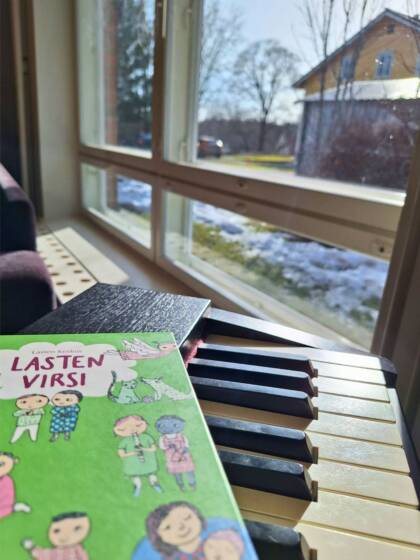 Lasten virsikirja kosketinsoittimen koskettimien päällä, takana ikkuna josta näkyy pihamaalle.