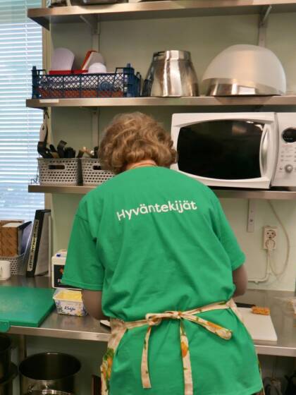 Nainen selin tekemässä ruokaa suurtalouskeittiössä ja hänen vihreässä t-paidassa lukee Hyväntekijä.