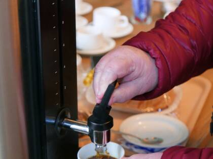Iäkkään henkilön kädet ottamassa kahvia termossäiliöstä ja taustalla kahvilan tiski, jolla kahvikuppeja ja voileipiä.