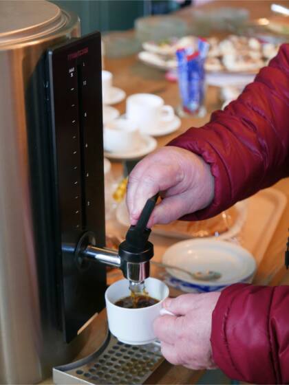 Iäkkään henkilön kädet ottamassa kahvia termossäiliöstä ja taustalla kahvilan tiski, jolla kahvikuppeja ja voileipiä.