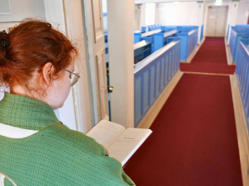 Nuori pappi vihreässä kasukassa katsoo kirjaa ja hänen taustallaan on kirkkosali punaisine käytävämattoineen sekä sinisine penkkeineen.
