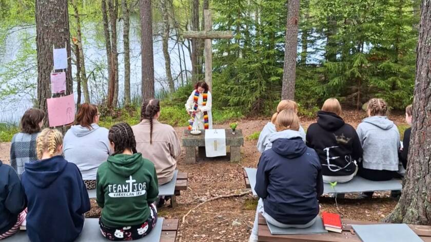 Nuoria istumassa penkeillä ulkona, järven metsäisellä rannalla ja heidän edessään on kivinen alttari, puinen risti ja nuori pappi valkoisessa albassa ja värikkäässä stolassa.
