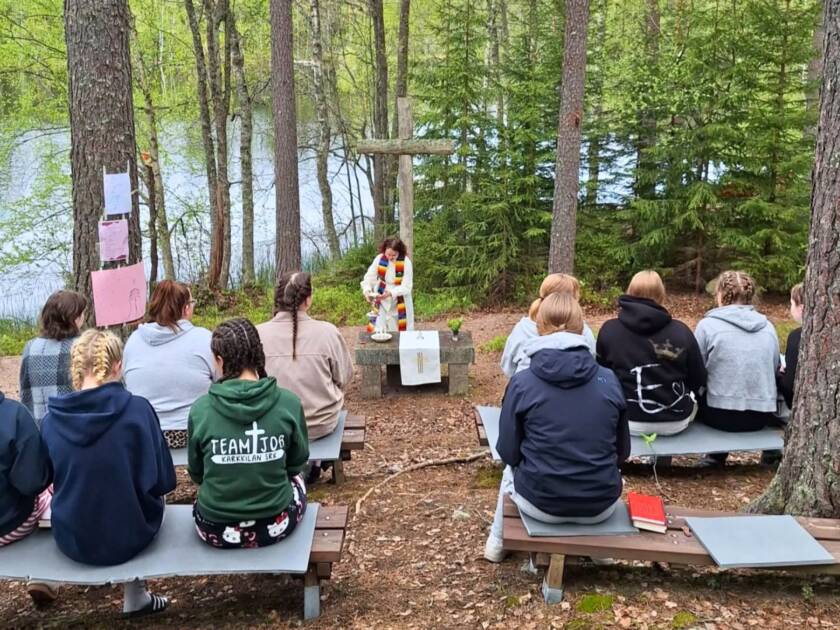 Nuoria istumassa penkeillä ulkona, järven metsäisellä rannalla ja heidän edessään on kivinen alttari, puinen risti ja nuori pappi valkoisessa albassa ja värikkäässä stolassa.