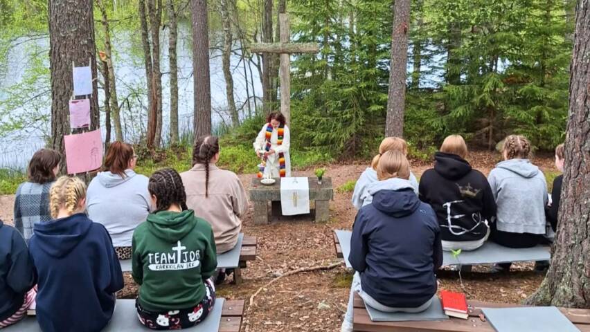 Nuoria istumassa penkeillä ulkona, järven metsäisellä rannalla ja heidän edessään on kivinen alttari, puinen risti ja nuori pappi valkoisessa albassa ja värikkäässä stolassa.