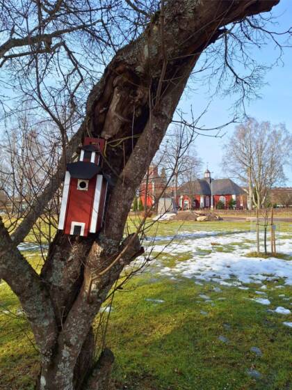 Etualalla puussa kiinni punainen, Karkkilan kirkkoa jäljittelevä linnunpönttö ja taustalla itse kirkko, sinistä taivasta ja nurmikkoa, jolla vähän lunta.