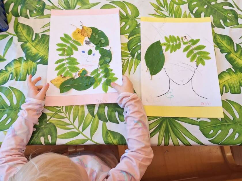 Pieni lapsi pitää kädessään piirtämällä ja vihreistä sekä keltaisista puun lehdistä tehtyä kuvaa ihmisen kasvoista. Vieressä toinen kuva.