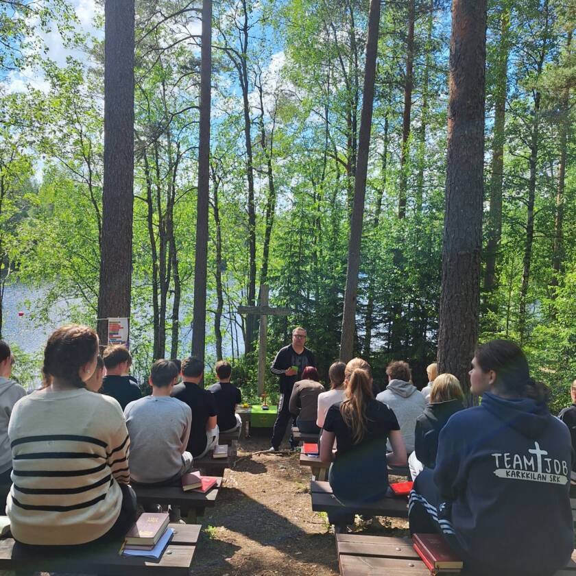 Nuoria istumassa selin aurinkoisena päivänä ulkona penkeillä ja edessä risti, alttari sekä mies puhumassa takanaan puita ja järvi.
